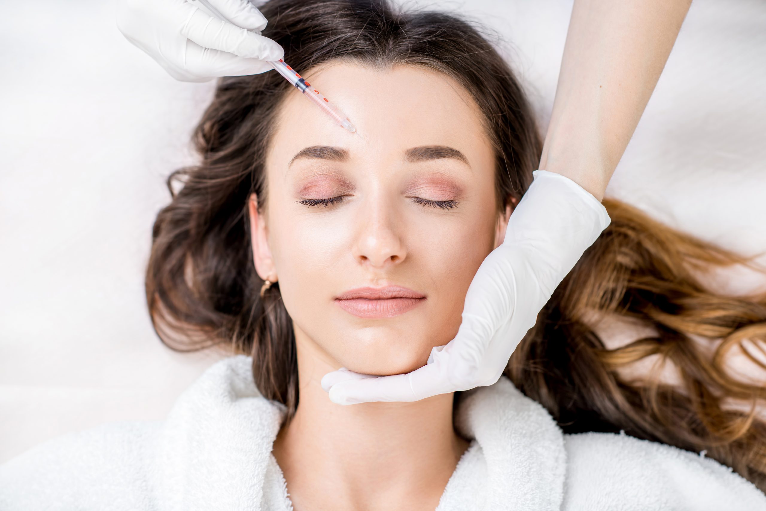 Woman receiving a botox injection in the eye zone lying in bathrobe on the medical couch