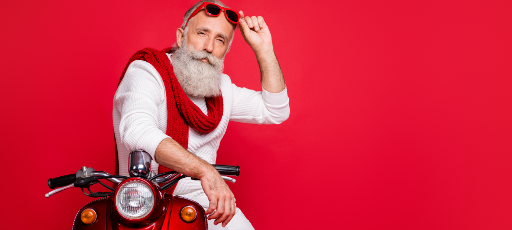Santa sitting on a scooter with sunglasses on his head against red backdrop